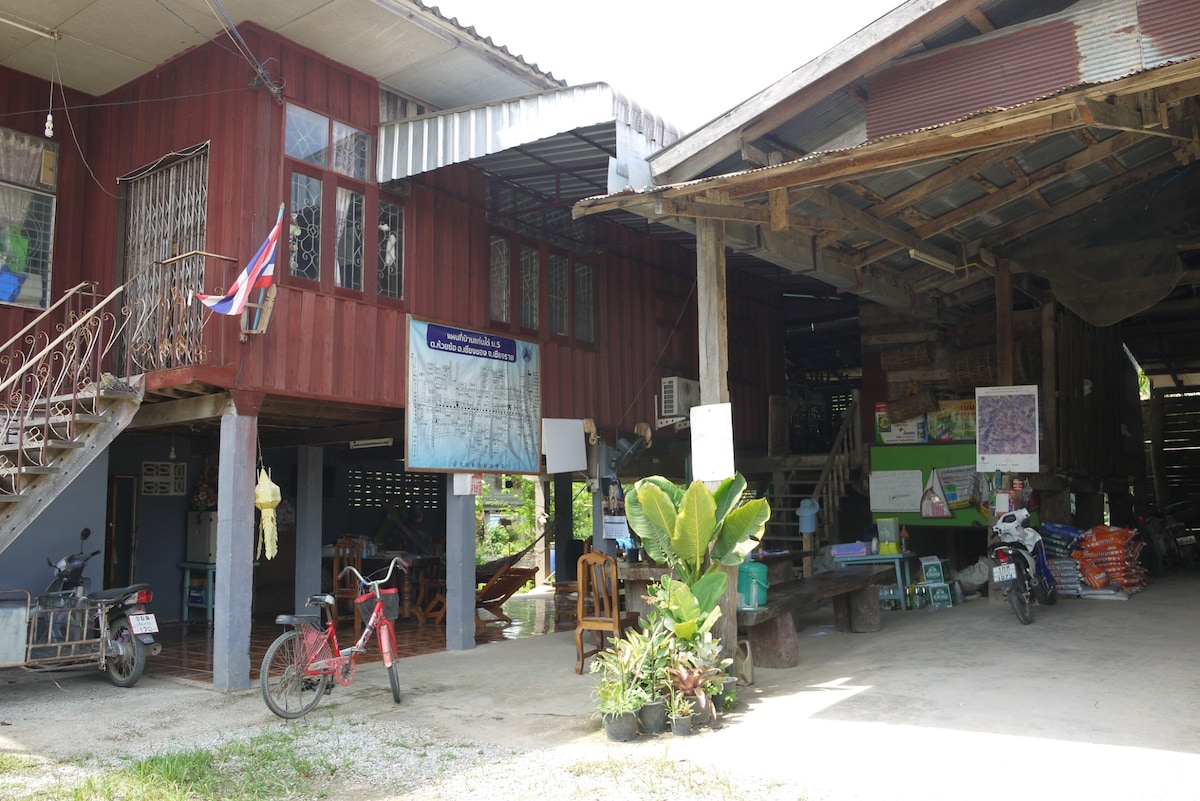 A traditional village building is depicted, featuring wooden exterior with open spaces. Bicycles and seating areas are visible outside, along with potted plants. Signage indicates local information, while the ground is paved, providing accessible paths for visitors.