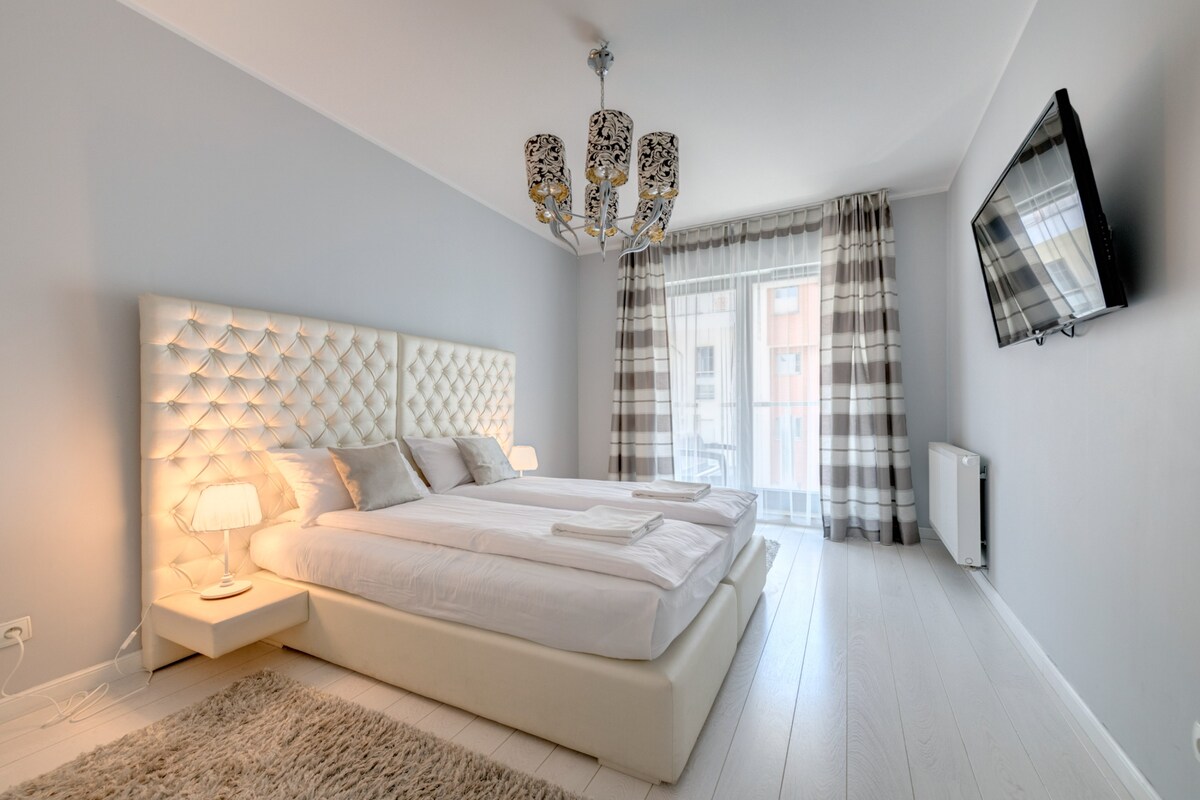 A well-appointed bedroom features a spacious bed with a tufted headboard, complemented by soft bedding. Two bedside lamps provide gentle lighting, while a modern chandelier adds elegance. A large window with sheer curtains lets in natural light, and a wall-mounted television is visible.