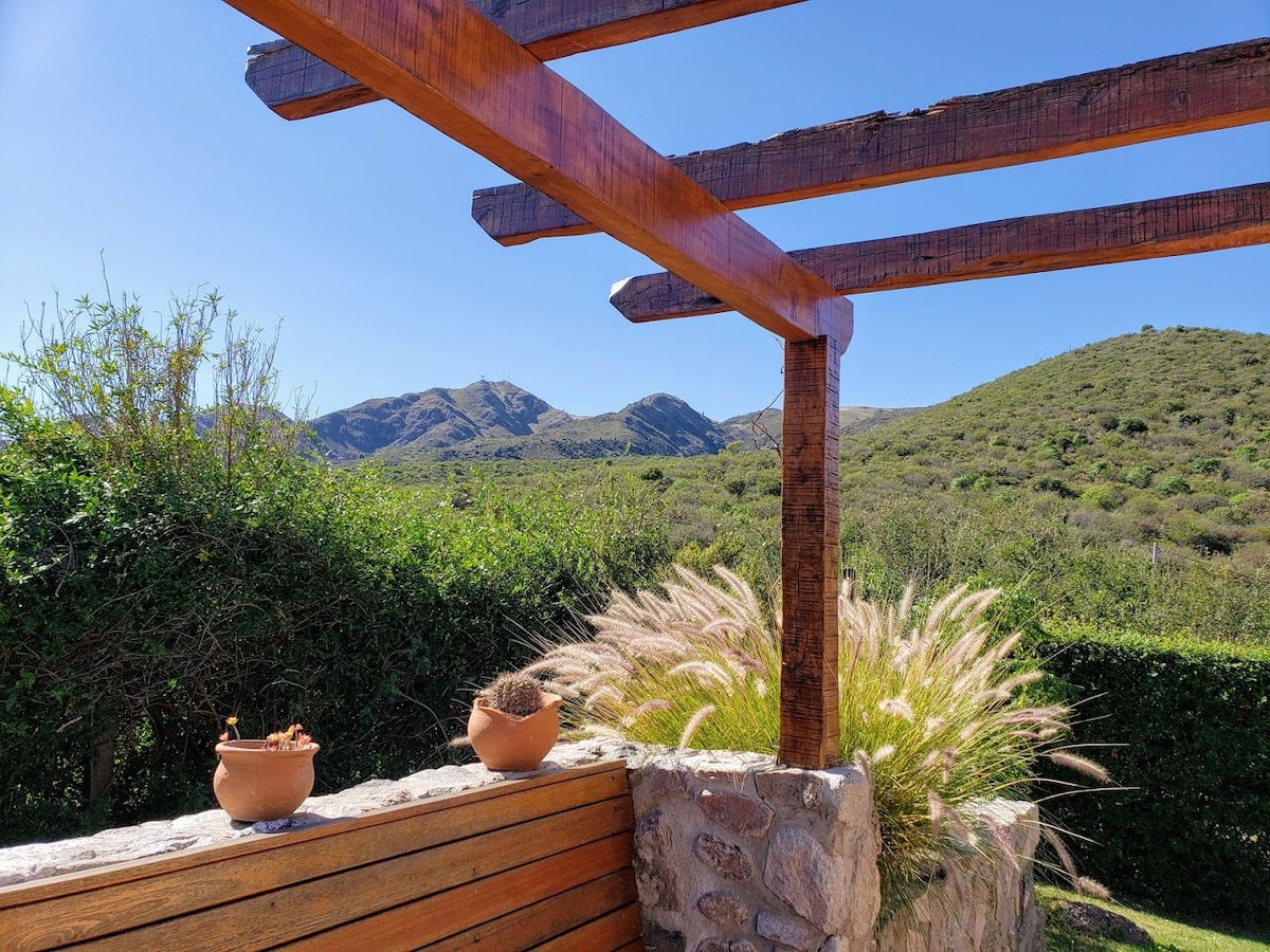 A wooden pergola frames a view of rolling green hills and distant mountains under a clear blue sky. Potted plants adorn the stone wall, and ornamental grasses softly sway, adding texture to the serene outdoor space.