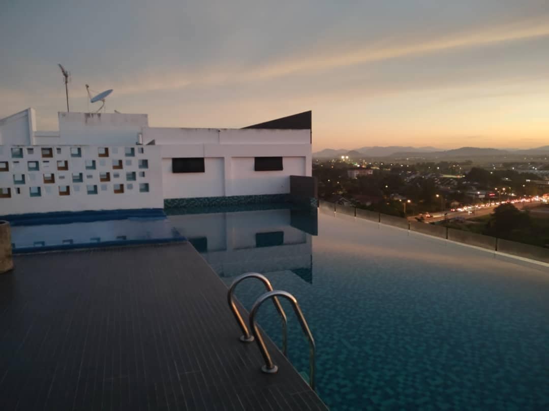 An infinity pool reflects the evening sky, with a view of distant hills and city lights. The pool's edge creates a seamless transition to the horizon, while three metal handrails provide access. The surrounding area showcases modern architectural elements against a tranquil backdrop.