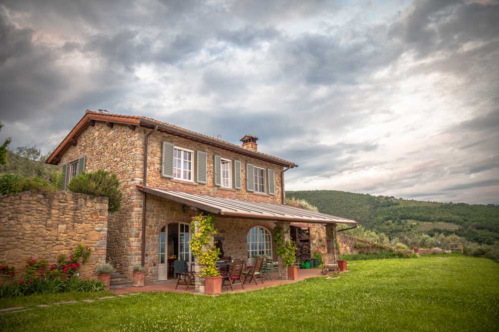 A charming stone villa is set against a scenic backdrop of lush green hills. The exterior features large windows and an inviting porch area with outdoor seating, surrounded by colorful garden plants, creating a peaceful and natural setting.
