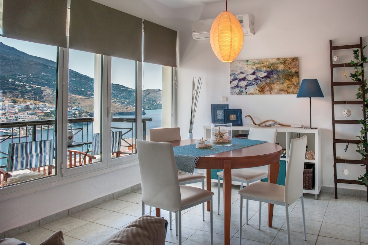 A dining area is highlighted by a round wooden table with a blue table runner, surrounded by four light-colored chairs. Large windows provide a panoramic view of the Aegean Sea and Batsi bay, with gentle natural light illuminating the space. A decorative lamp and wall art add to the ambiance.