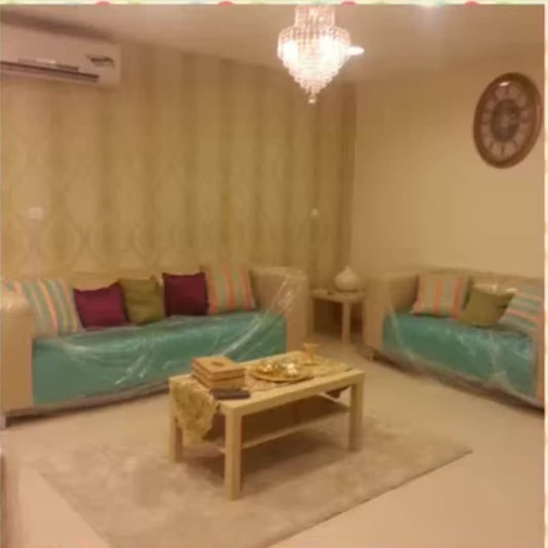 A comfortable living area is highlighted by two sofas adorned with colorful cushions. A coffee table sits in the center, complemented by decorative items. A soft rug offers warmth underfoot, while a chandelier provides gentle illumination, creating a cozy environment.