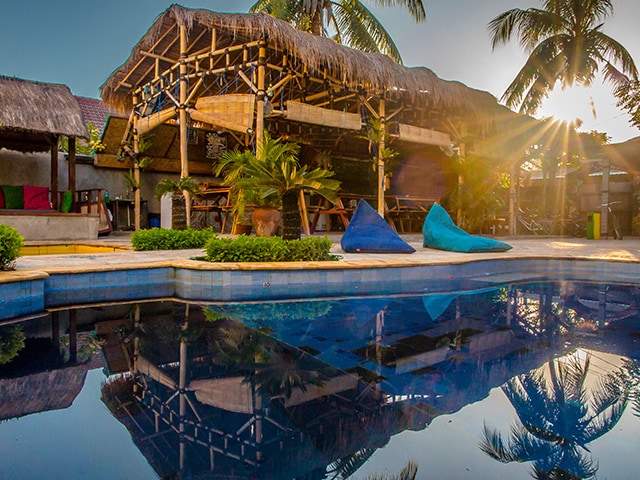 A spacious communal area features a thatched roof structure surrounded by palm trees and vibrant outdoor furniture. The tranquil swimming pool reflects the natural beauty, while sunlight filters through, creating a serene ambiance for guests to enjoy.