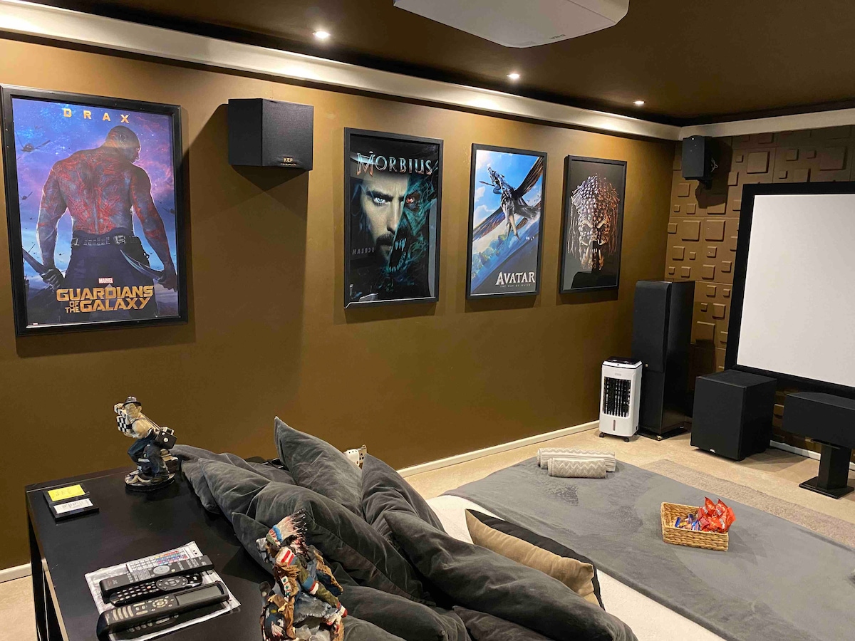 A cozy home cinema room is shown, featuring an inviting seating area with plush cushions. Movie posters hang on a warm-toned wall, while a large projection screen is positioned prominently. The space is designed for an immersive viewing experience, with sound speakers placed strategically.
