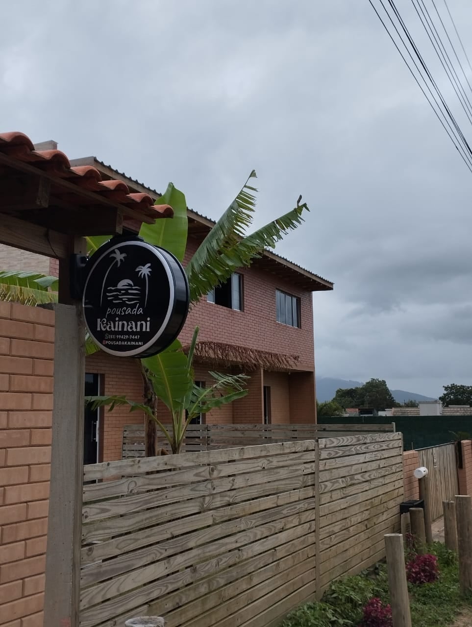 A sign displaying the name 'Pousada Kainani' is mounted on a wooden fence, framed by lush green banana plants. The building with a terracotta roof reflects a modern design, complemented by cloudy skies in the background.