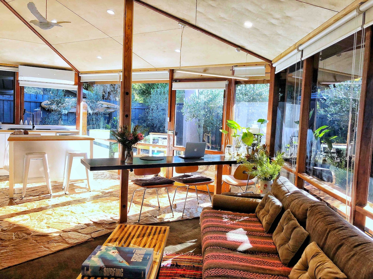An open-concept living space is framed by large windows, allowing natural light to fill the area. A cozy sectional sofa is positioned alongside a wooden coffee table. A modern dining table is visible, accompanied by minimalistic seating, while lush greenery can be seen through the transparent panels.