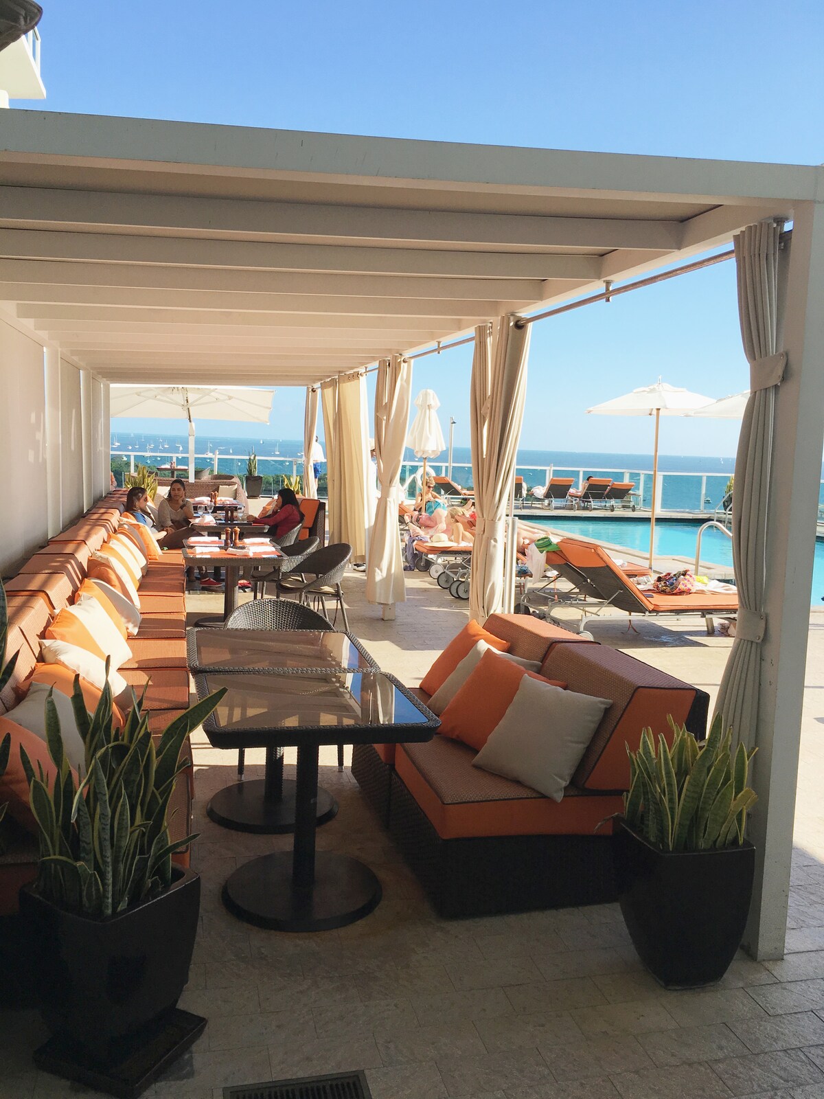A comfortable outdoor seating area is shaded by a canopy, featuring plush orange and beige cushions on a lengthy sofa. A glass table sits in front, surrounded by potted plants. In the background, lounge chairs and umbrellas line the edge of a sparkling pool, with expansive views of the sky.