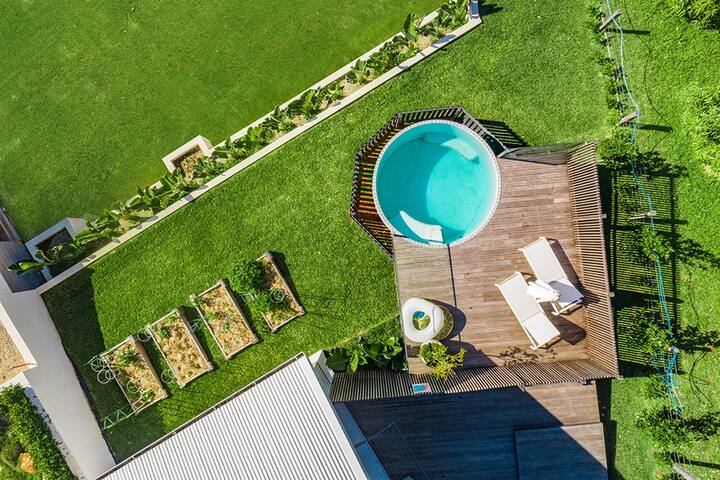 An aerial view of a backyard featuring a circular above-ground pool with a surrounding wooden deck and two lounge chairs. The lawn includes a small garden with raised beds and a row of neatly arranged plants along the edge of the yard
