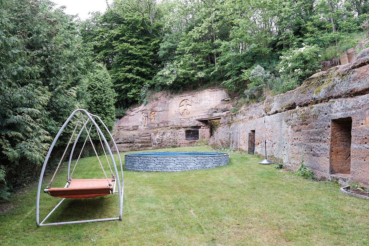 A serene outdoor area is featured, showcasing a circular stone pool surrounded by lush greenery. A unique swing chair is positioned nearby, offering a place to relax. Natural rock formations serve as a backdrop, enhancing the tranquil setting.