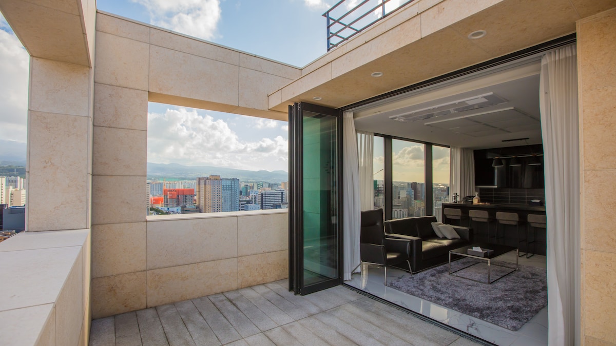A spacious terrace is accessible from the open living area, featuring modern seating arrangements and views of the city skyline. Large glass doors allow natural light to flood the interior, with minimalist decor and a plush area rug enhancing the space.