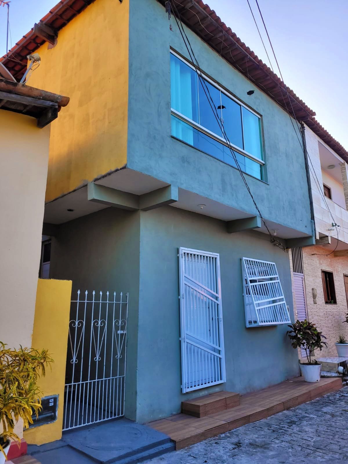 A two-story building is depicted, featuring a contemporary facade with a large window on the second floor. A white gate secures the entrance, complemented by wooden steps leading up to the door. The surroundings include colorful neighboring buildings.