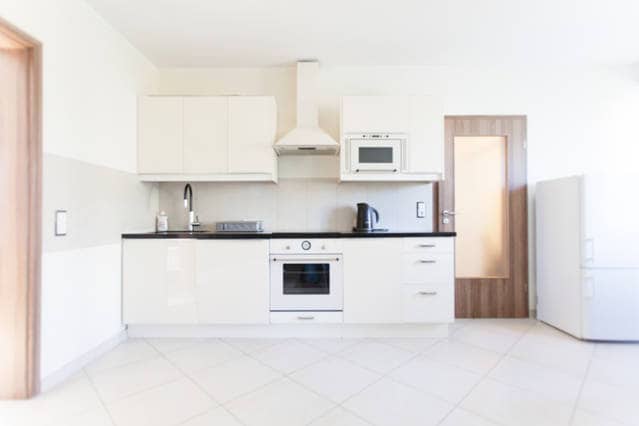 A modern kitchen features sleek, white cabinetry paired with a black countertop. Appliances include a built-in oven, microwave, and a kettle. A refrigerator is visible in the corner. The space is well-lit with neutral-colored walls and tiled flooring.