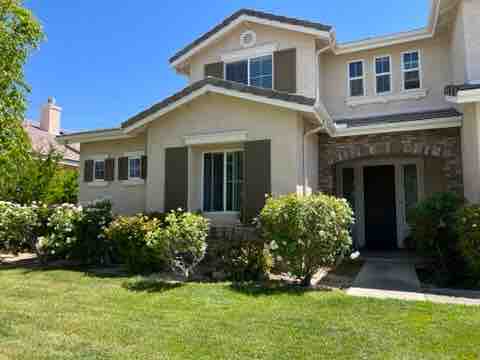 The exterior of the casita is showcased, featuring a well-maintained facade with light-colored stucco and decorative shutters. Lush greenery surrounds the entrance, complemented by a neatly manicured lawn. Clear blue skies offer a bright backdrop, enhancing the serene setting of the neighborhood.
