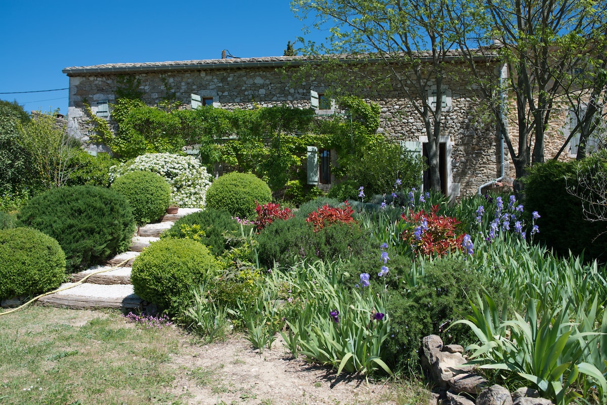 A charming stone house is framed by a well-maintained garden featuring various shrubs and colorful flowers. Pathways lined with greenery lead to the entrance, enhancing the tranquil ambiance of the space.