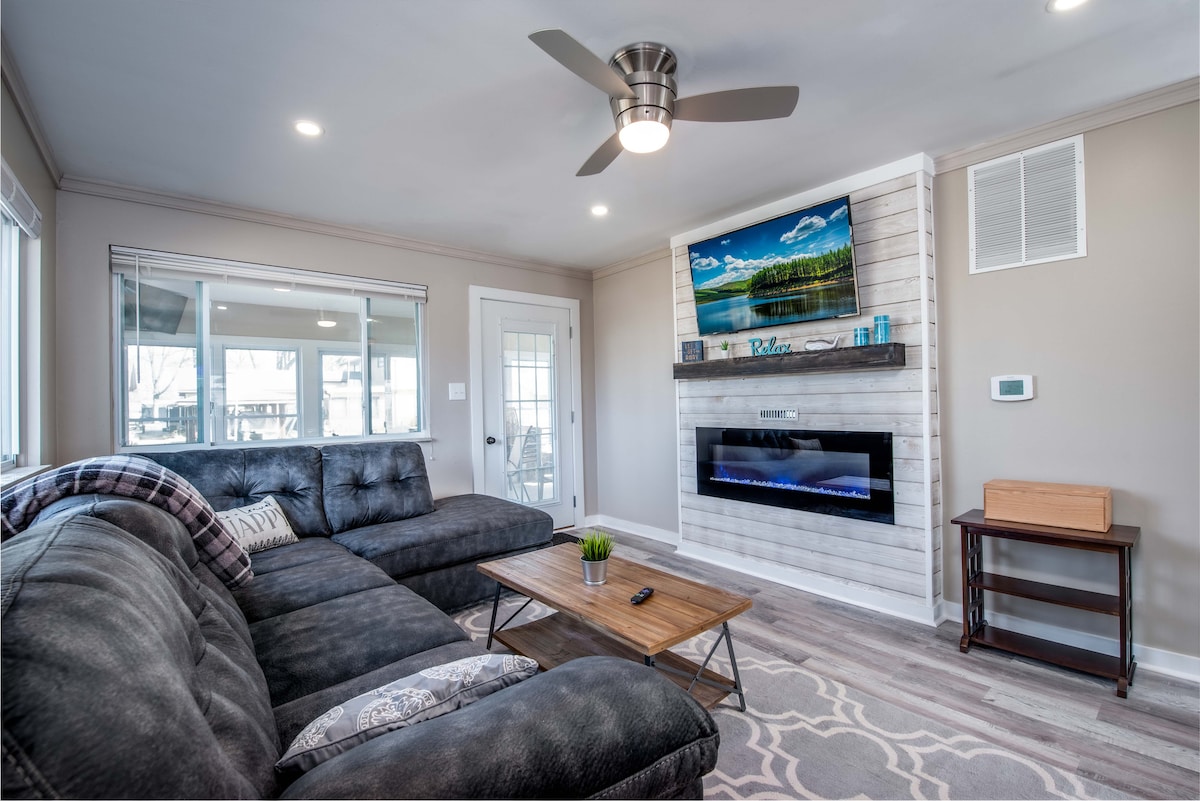 A living area features a plush sectional sofa with a textured finish and a wooden coffee table. A modern television is mounted above an electric fireplace on a paneled wall, while large windows provide natural light and views of the surroundings.
