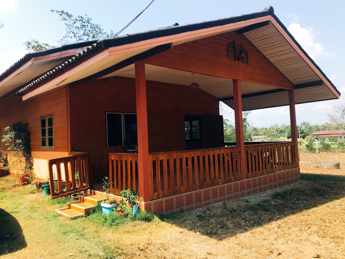 A wooden house with a spacious front porch stands surrounded by natural greenery. The porch features a railing and steps leading to the entry. Windows are positioned on each side, providing views of the outdoor landscape under a clear sky.