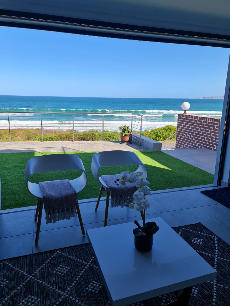 An open living area features a pair of white chairs with blankets draped over them. A small table holds a potted plant. Large sliding doors provide an unobstructed view of the ocean and beach, enhancing the connection between indoor and outdoor spaces.