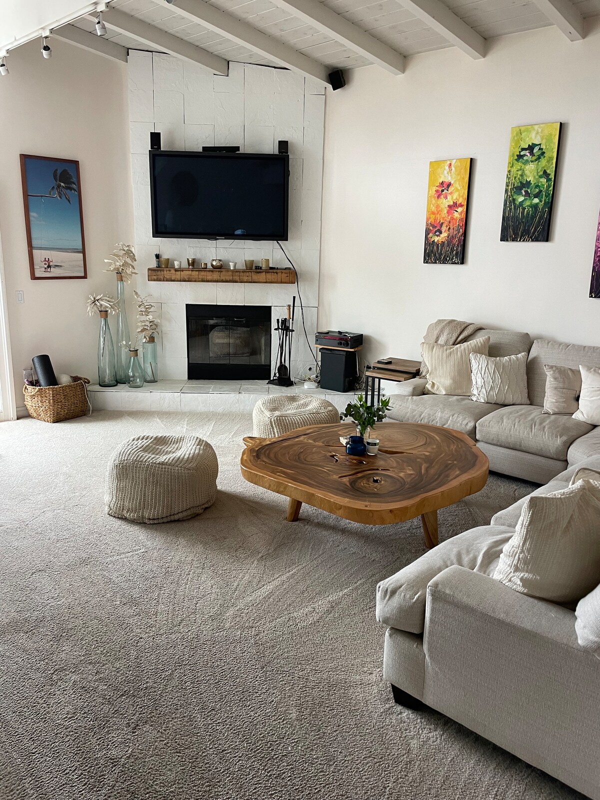 A spacious living area is highlighted by a large, natural wood coffee table and a cozy sectional sofa. The room features a fireplace and a mounted TV, alongside wall art in vibrant colors. Natural light enters through a nearby window, enhancing the inviting space.