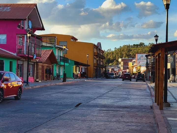A vibrant street scene reveals colorful buildings featuring various architectural styles. The wide, smooth road invites exploration, while street lamps line the sides, providing illumination. Vehicles are parked along the curb, and a backdrop of greenery can be seen in the distance.