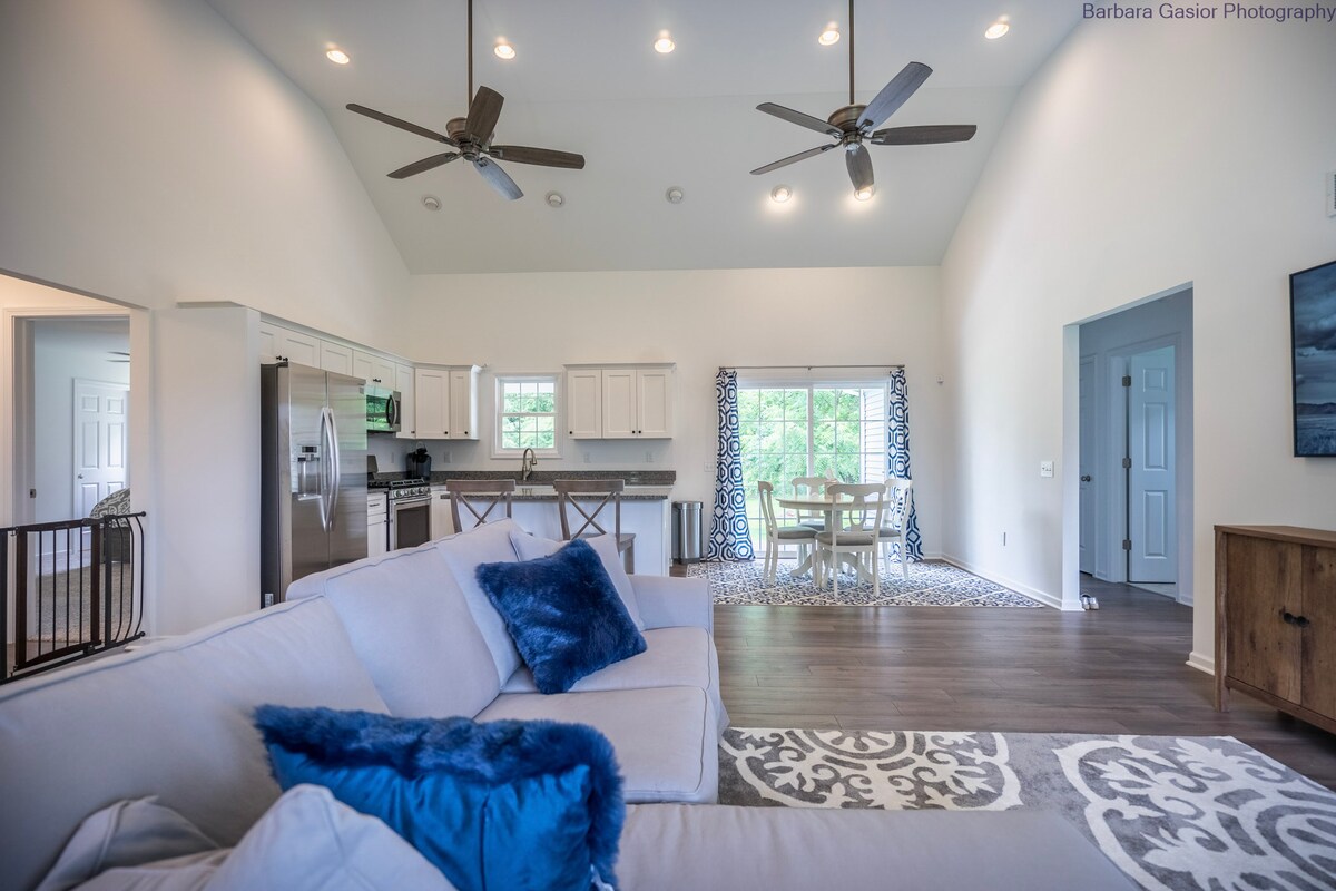 A spacious living area features high vaulted ceilings and multiple ceiling fans. Light-colored cabinetry and stainless steel appliances are visible in the kitchen. A dining table with chairs is situated near large windows framed with patterned curtains, allowing natural light to enhance the room.