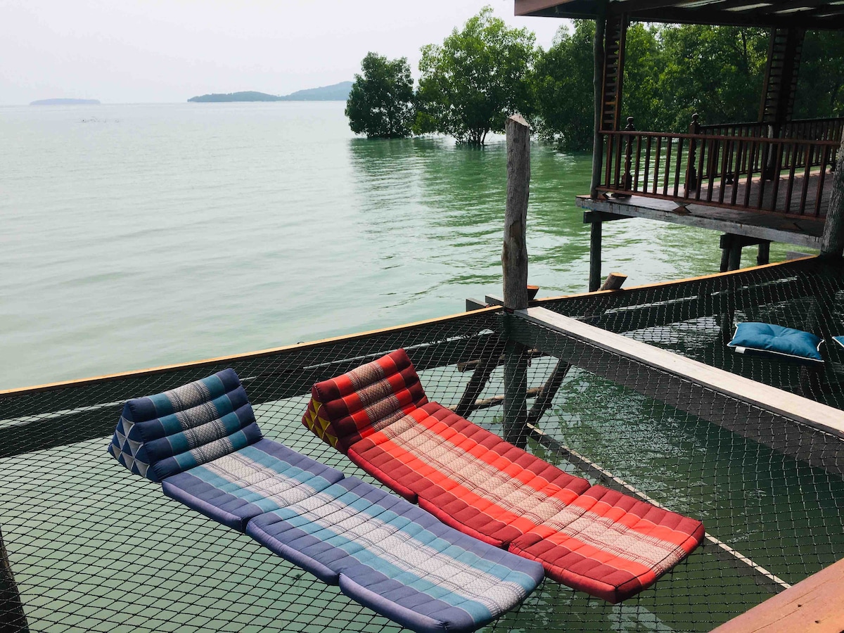 Two brightly colored mats are positioned on a net over water, providing a relaxed space for lounging. Surrounding greenery is visible in the background, enhancing the tranquil setting. The structure features wooden elements, blending seamlessly with the natural surroundings.