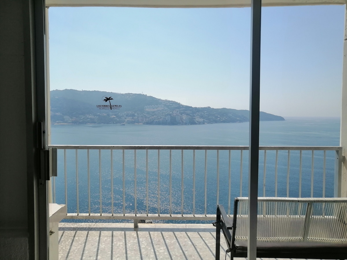 A serene balcony view is captured, showcasing a panoramic glimpse of the ocean and distant coastline. The calm waters reflect daylight, while the horizon blends seamlessly into the clear sky. A minimalist railing and bench are visible, enhancing the tranquil scene.