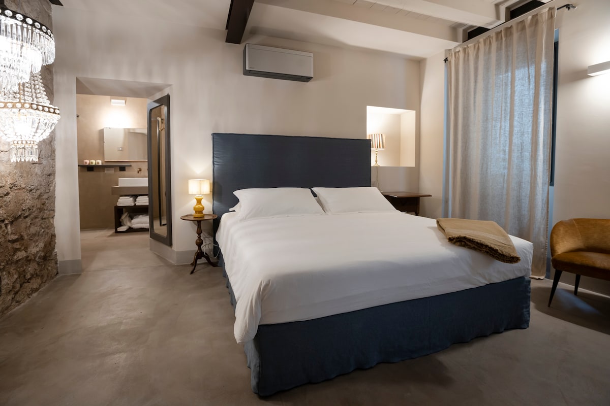 A spacious room features a plush bed with crisp white linens, complemented by gentle lighting from a bedside lamp. A large window dressed with sheer curtains allows natural light to enter, and a cozy armchair adds a welcoming touch to the space.