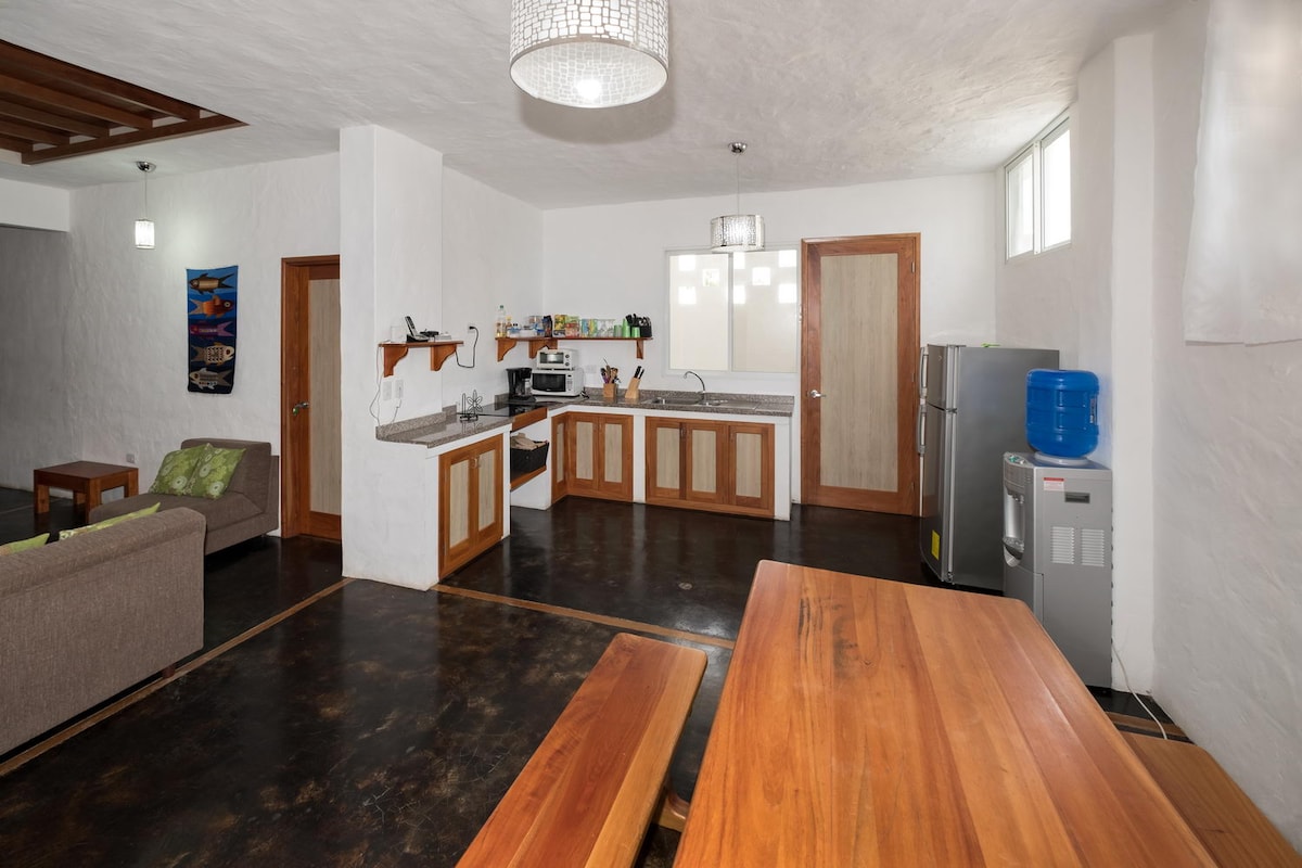 A spacious kitchen area with wooden cabinetry and a modern refrigerator is visible. A dining table made of light wood is set in the foreground. A comfortable seating area featuring a sofa is positioned to the left, complemented by natural light from nearby windows.