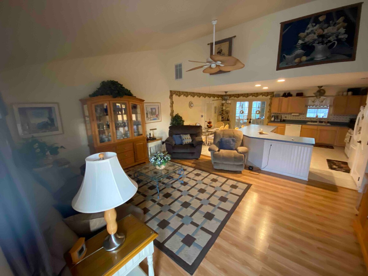 An open living area is featured, highlighted by hardwood flooring and a large area rug. Two comfortable seating arrangements are positioned near a dining space. Natural light flows through the windows, illuminating decorative elements including plants and framed artwork on the walls.