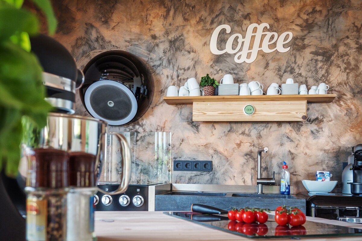 A modern kitchen space features a wooden shelf displaying white mugs and a decorative 'Coffee' sign. Fresh tomatoes are placed on the countertop, alongside a coffee grinder and various kitchen appliances. Textured walls in earthy tones add character to the room.