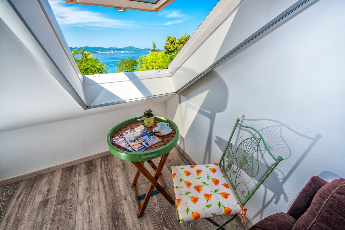 A cozy corner captures a charming table with a small plant, paired with a patterned chair. Sunlight streams through a skylight, illuminating the space and offering a view of the sea and greenery beyond.
