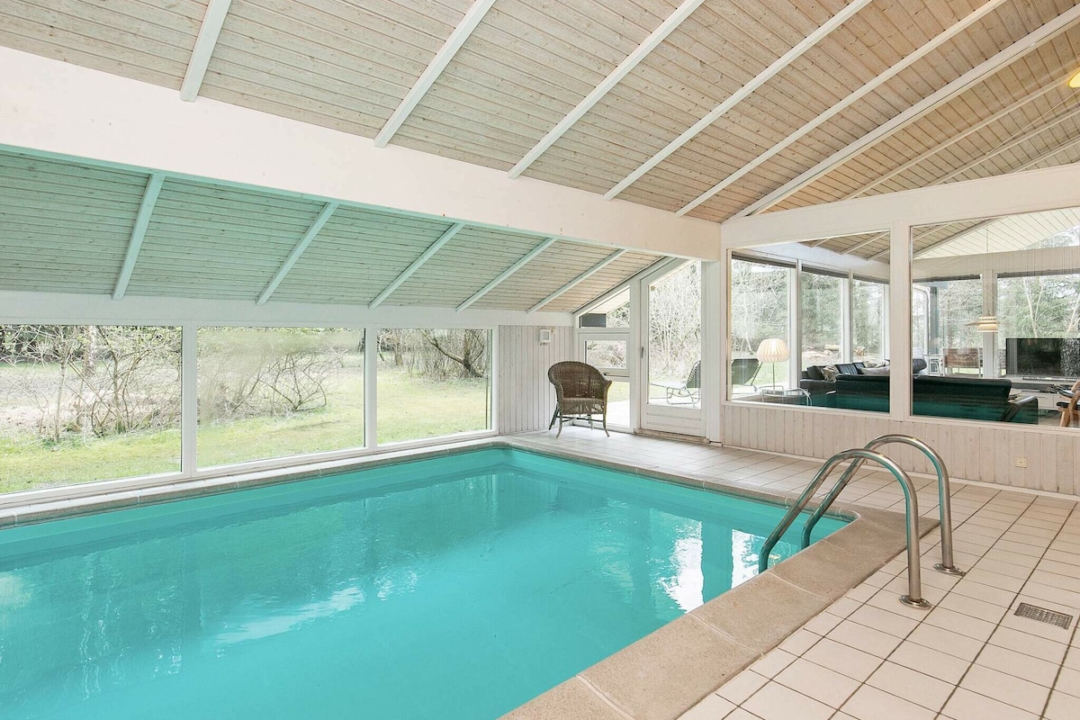 An indoor pool is framed by large windows, allowing natural light to illuminate the space. The pool features a tiled border and a gentle entry with steps. A cozy seating area is visible beside the pool, enhancing the relaxation potential of the environment.