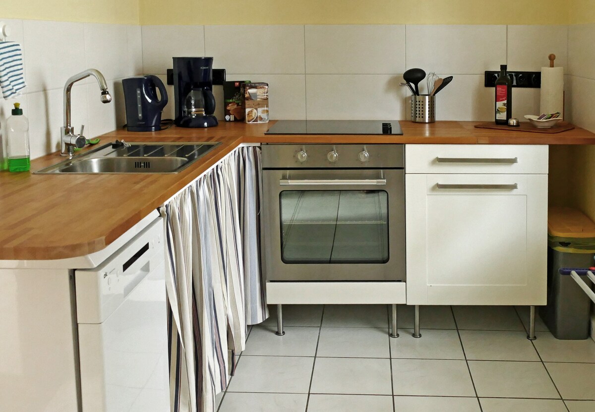 A well-equipped kitchen features a stainless steel oven and a cooktop integrated into a wooden countertop. A dishwashing machine is present, alongside a coffee maker and kitchen utensils organized in a holder. Light-colored tiles cover the floor, complemented by a striped curtain under the sink.