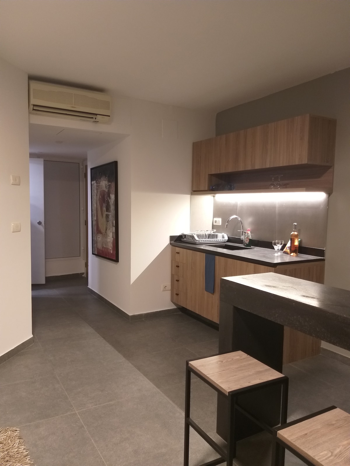 A modern kitchenette is featured with wooden cabinetry and a dark countertop. Glassware is neatly arranged next to a sink, while a small bar table with stools adds functionality. Soft lighting enhances the workspace, creating a clean and organized atmosphere.