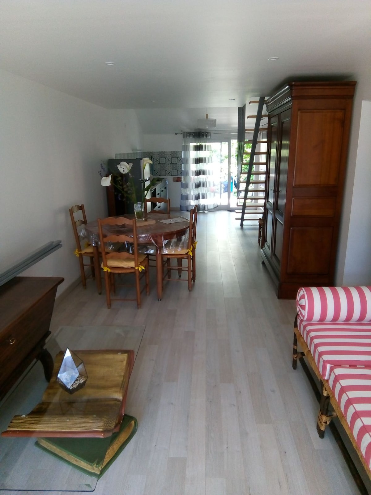 A bright and open living space features light-colored wooden flooring and a mix of furnishings. A wooden dining table with chairs sits near the window, while a striped couch adds color to the room. Natural light flows through large glass doors leading to an outdoor area.