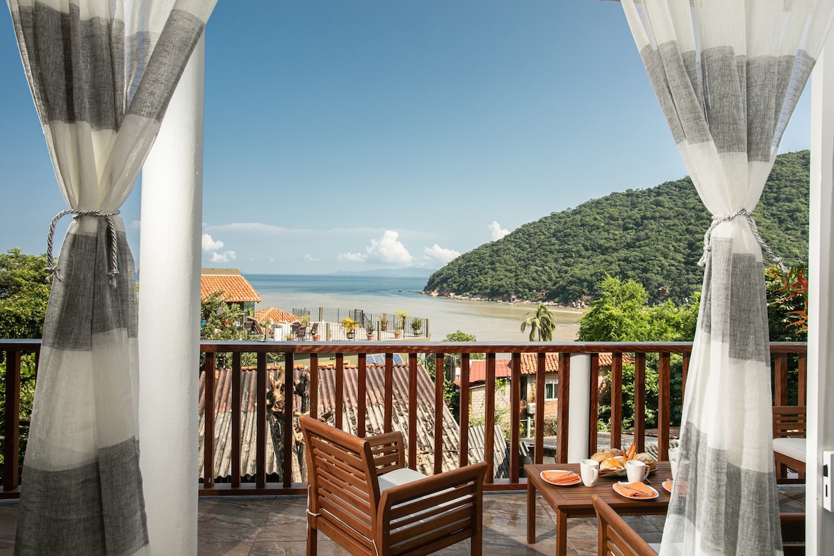 A balcony with light, sheer curtains offers a view of the ocean and lush hills. Two wooden chairs and a table are set with breakfast items, inviting relaxation while enjoying the surrounding natural beauty.