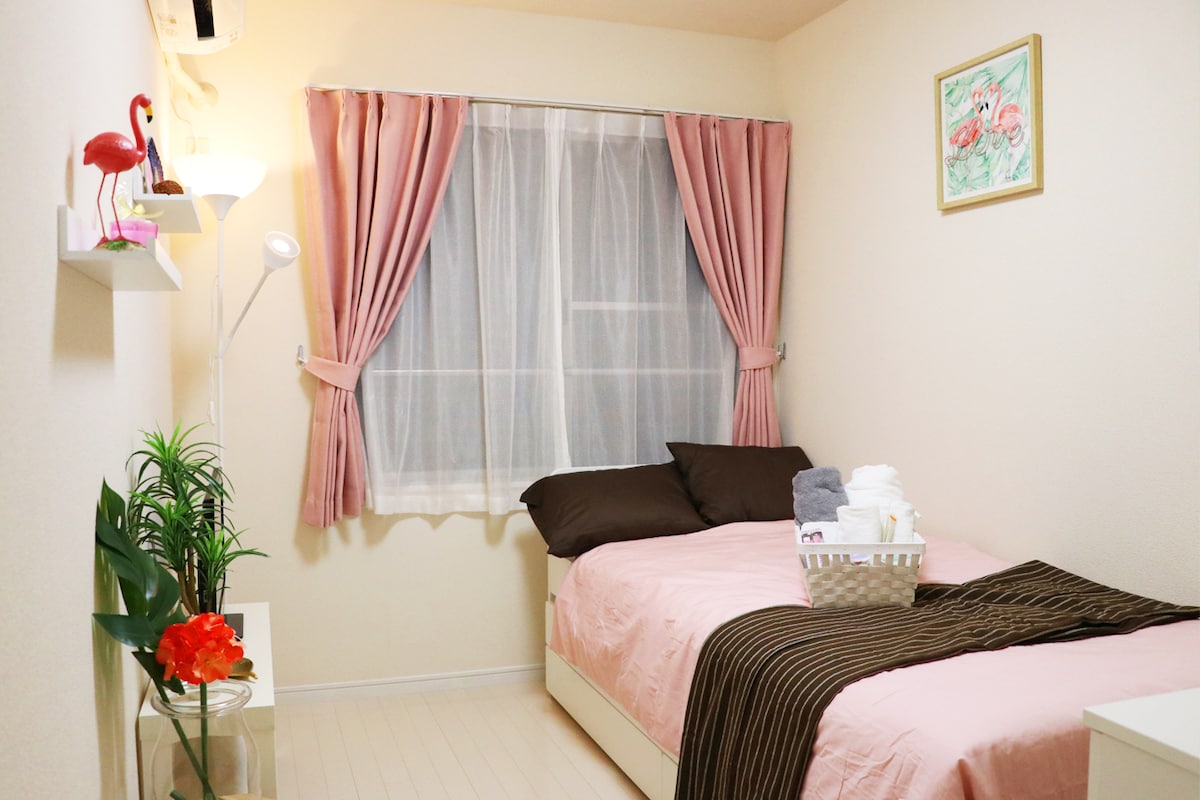 A cozy guest room features a single bed with pale pink bedding and a dark striped throw blanket. Soft curtains frame the window, and a small plant adds a touch of greenery. A basket of neatly arranged towels sits on the bed, complemented by a floral wall art piece.