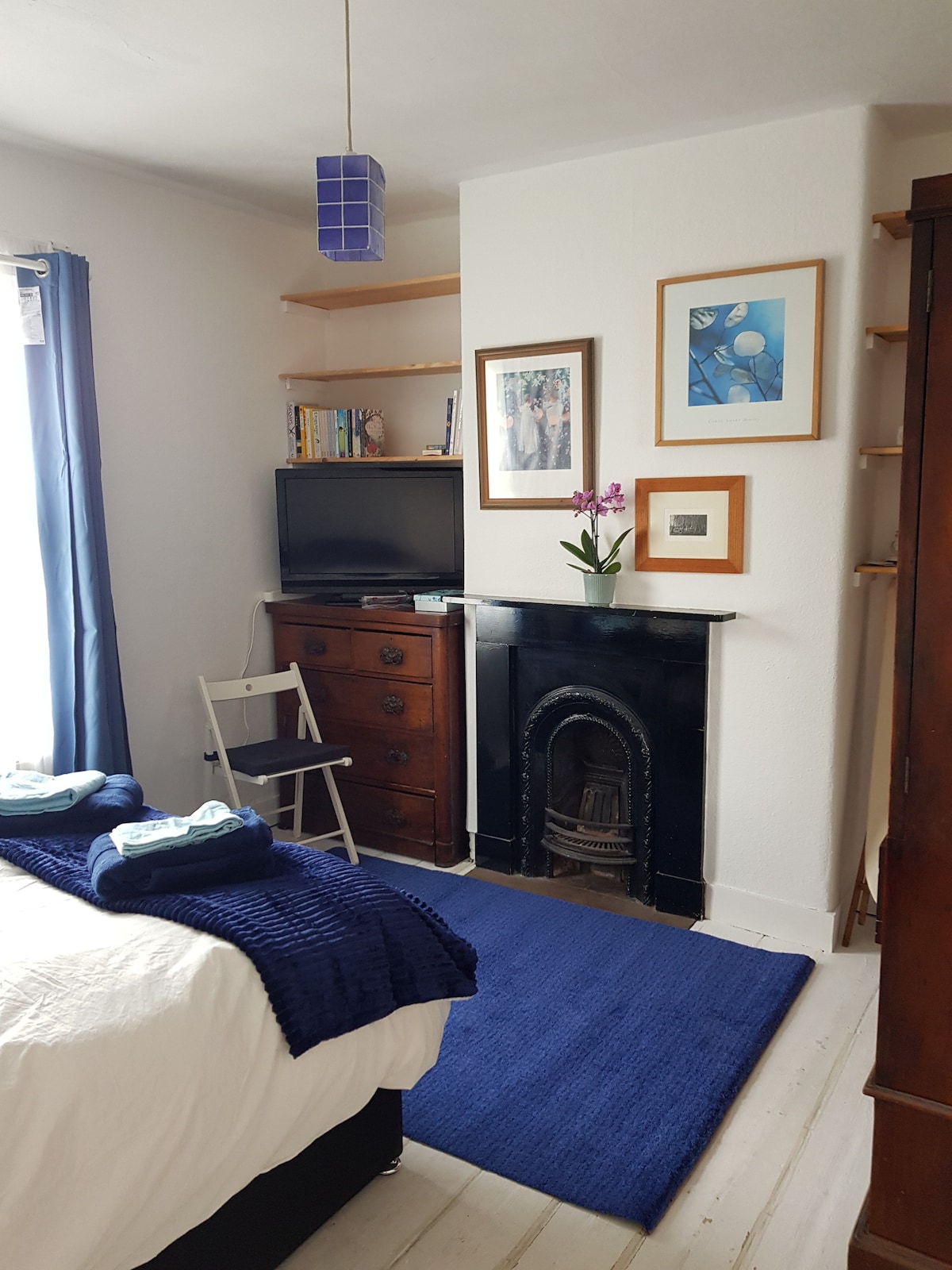 This cozy bedroom features a comfortable bed with neatly arranged linens. A small bookshelf and a wooden cabinet provide storage. A television rests on top of the cabinet, and framed artwork adorns the walls. A flower arrangement adds a touch of color to the room.