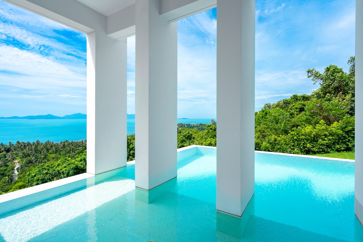 A serene infinity pool glistens in bright turquoise, extending towards the horizon. Tall white columns create an open space that frames the stunning sea view, surrounded by lush greenery and the distant outline of islands. The clear sky reflects on the water's surface.