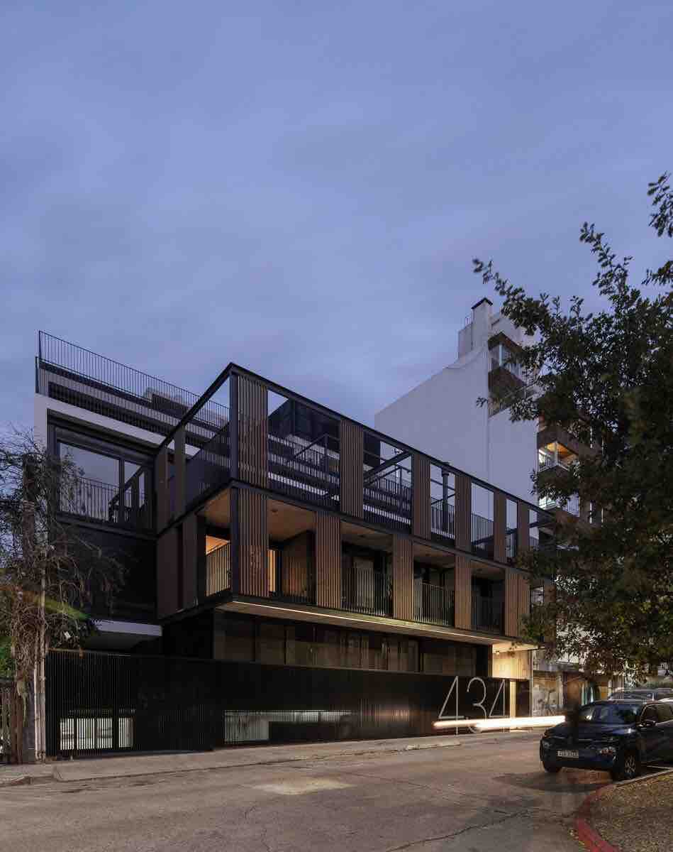 A contemporary building features a striking design with a combination of dark wooden panels and glass. Its multiple balconies extend outward, offering views of the surrounding area. The street is lined with trees, enhancing the urban landscape.