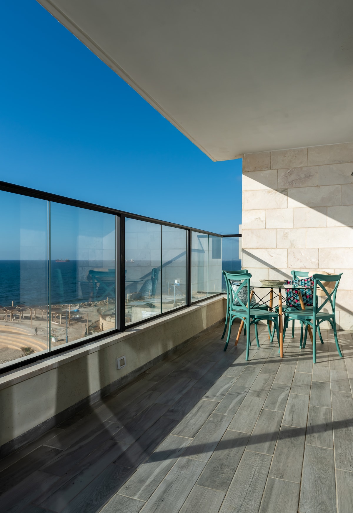 A balcony is displayed with a small dining table and four chairs. It features a glass railing that provides an unobstructed view of the Mediterranean Sea and surrounding landscape. The floor has a wooden finish, adding a modern touch to the outdoor space.