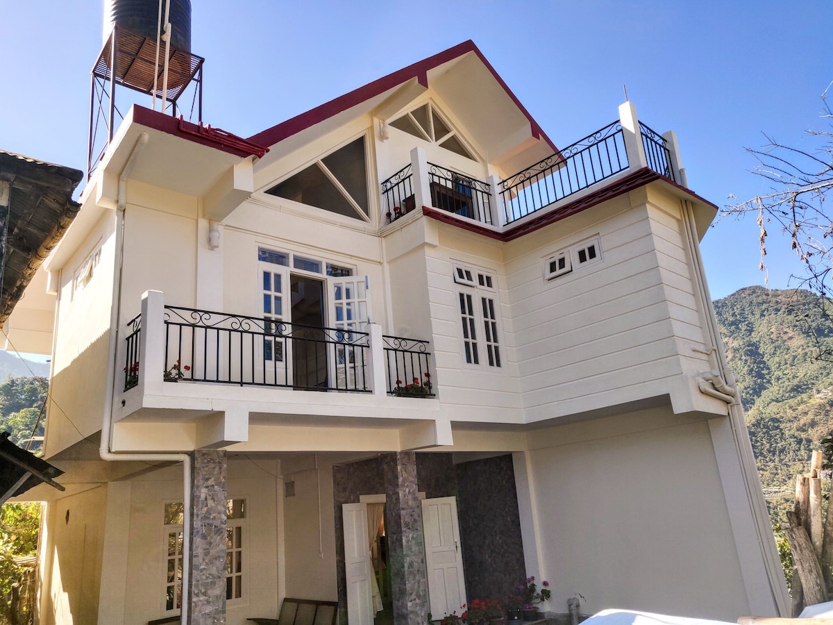 The two-story cottage features a modern design with a sloping red roof and expansive windows, complemented by a balcony on the upper level. Stone accents and a water tank on the rooftop add character. Surrounding mountains are visible in the background.