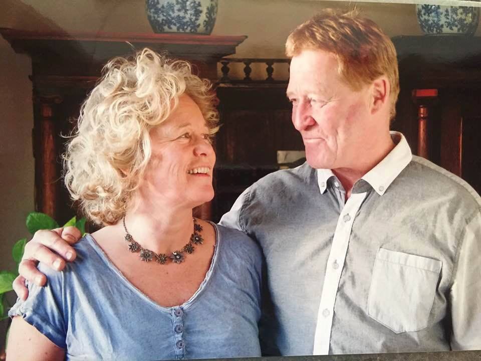 A close-up image features two individuals with cropped faces standing together. One person wears a light blue shirt and a decorative necklace, while the other is dressed in a light grey button-up shirt. A wooden backdrop with shelves is visible behind them, adorned with decorative items.
