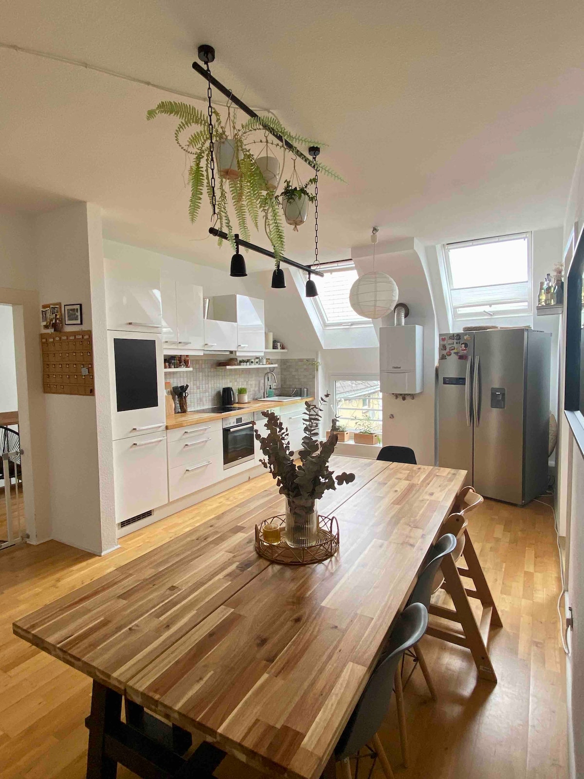The kitchen features a large wooden dining table surrounded by several chairs, offering a communal space. A variety of modern appliances are visible, including a refrigerator and a stove. Skylights bring natural light into the room, enhancing the fresh greenery above the table.