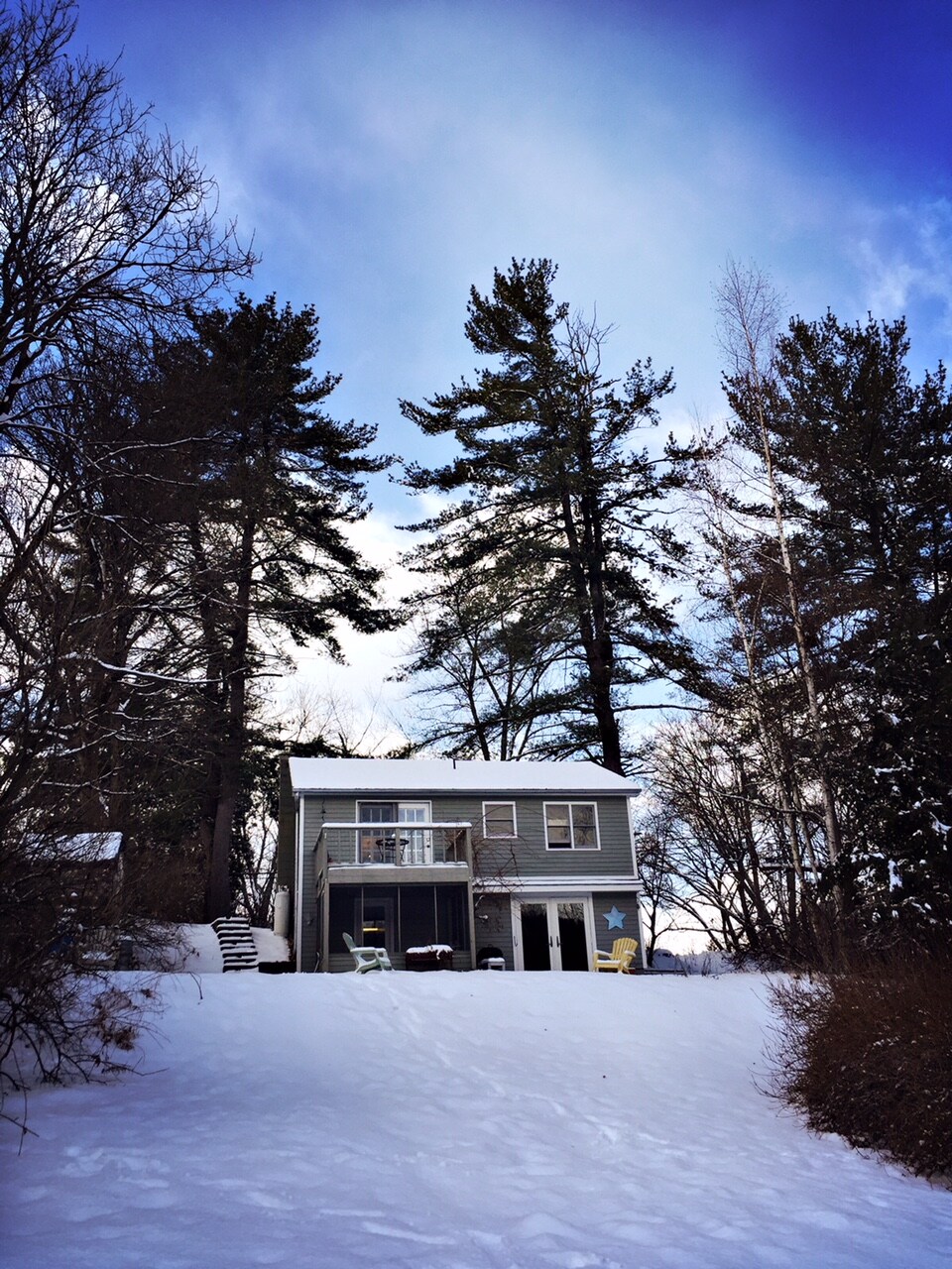 A two-story house sits atop a snowy slope, framed by tall pine trees. The exterior features a light grey façade with decorative elements, including a star. A balcony is seen above a ground-level entrance, with two colorful chairs positioned on the front porch.