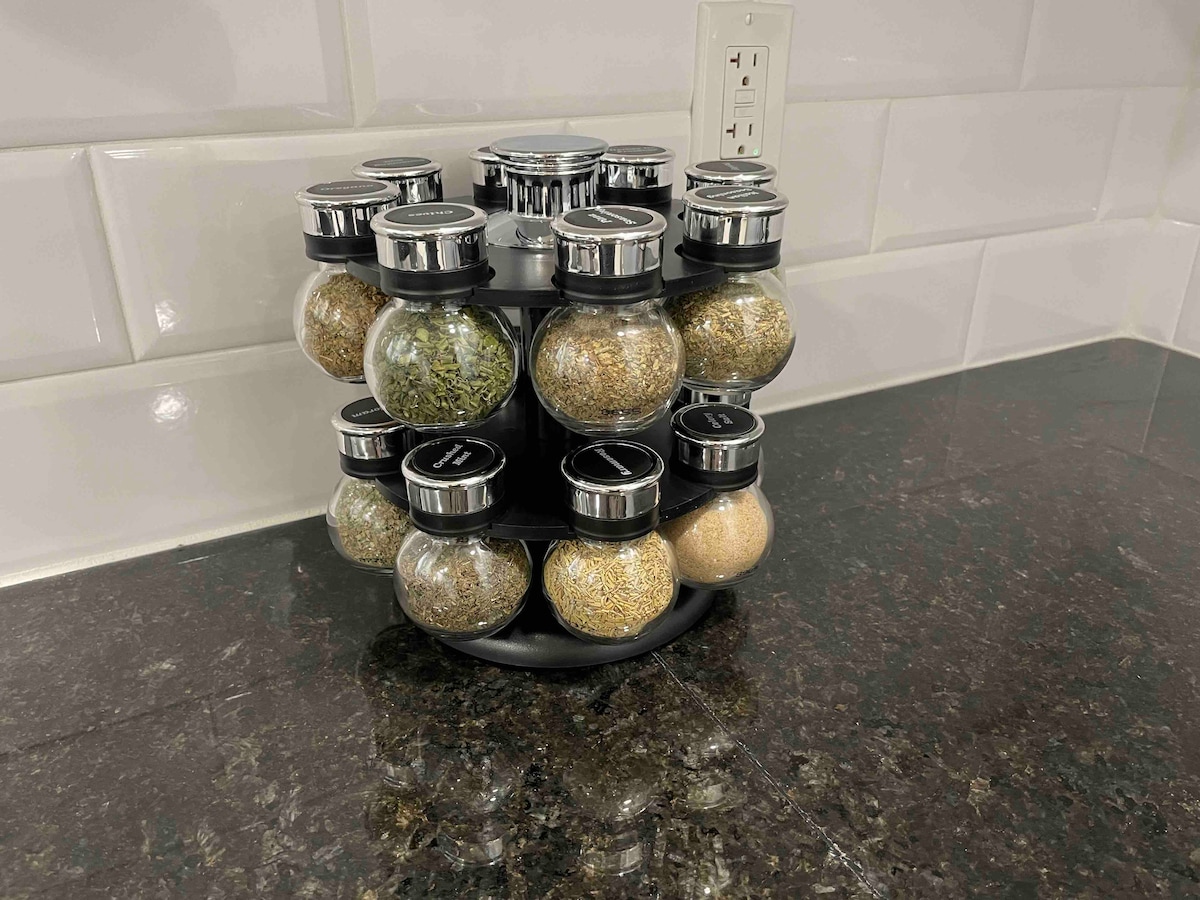 A rotating spice rack is depicted, featuring twelve glass jars filled with various spices and herbs. The jars have clear lids with black tops and are arranged in a circular stand. The spice rack is placed on a dark granite countertop, complemented by white subway tile in the background.