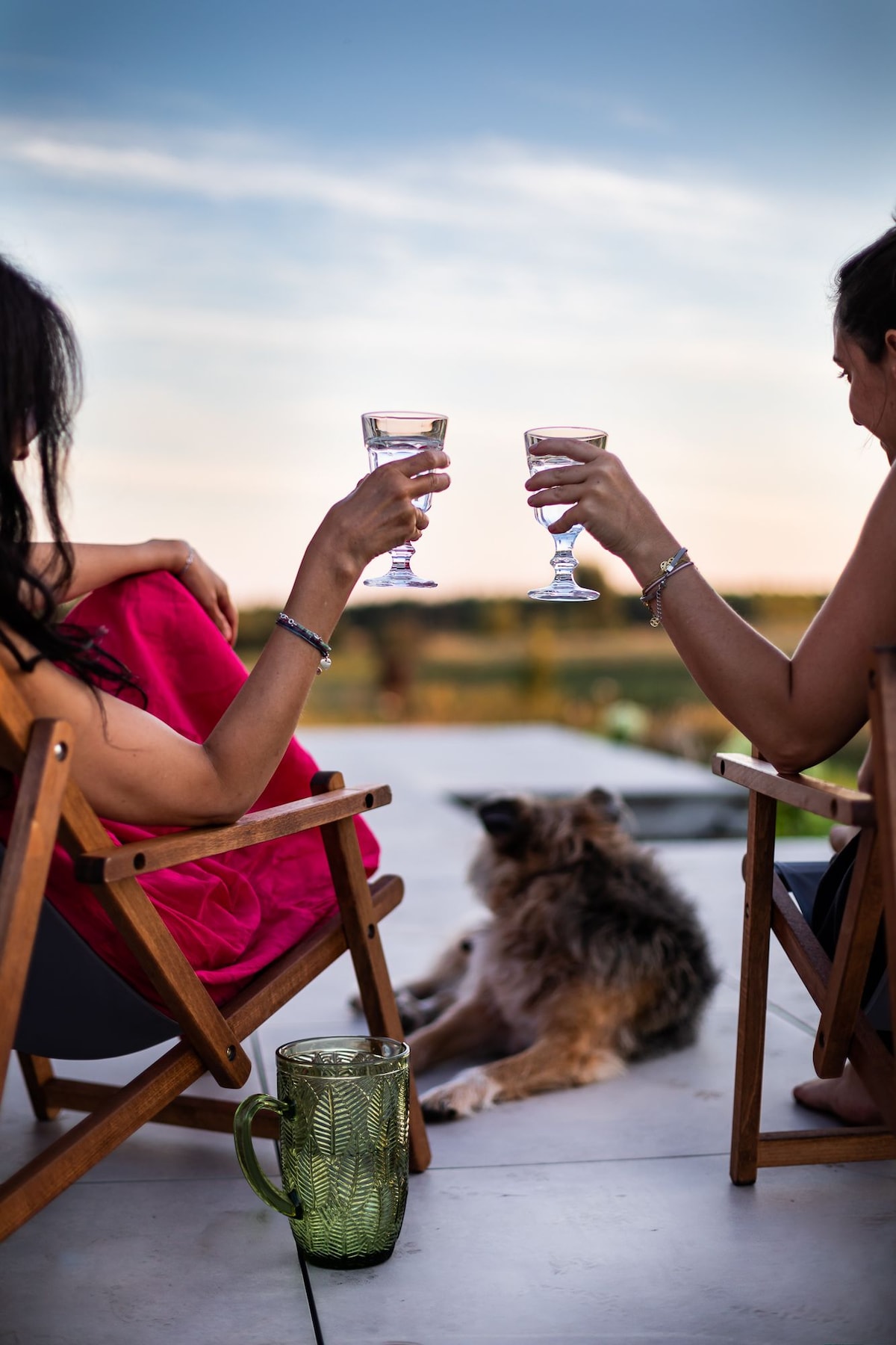Two individuals seated in wooden chairs enjoy drinks with a calm landscape in the background. A dog sits comfortably at their feet, contributing to a serene atmosphere as the sun sets over the horizon.