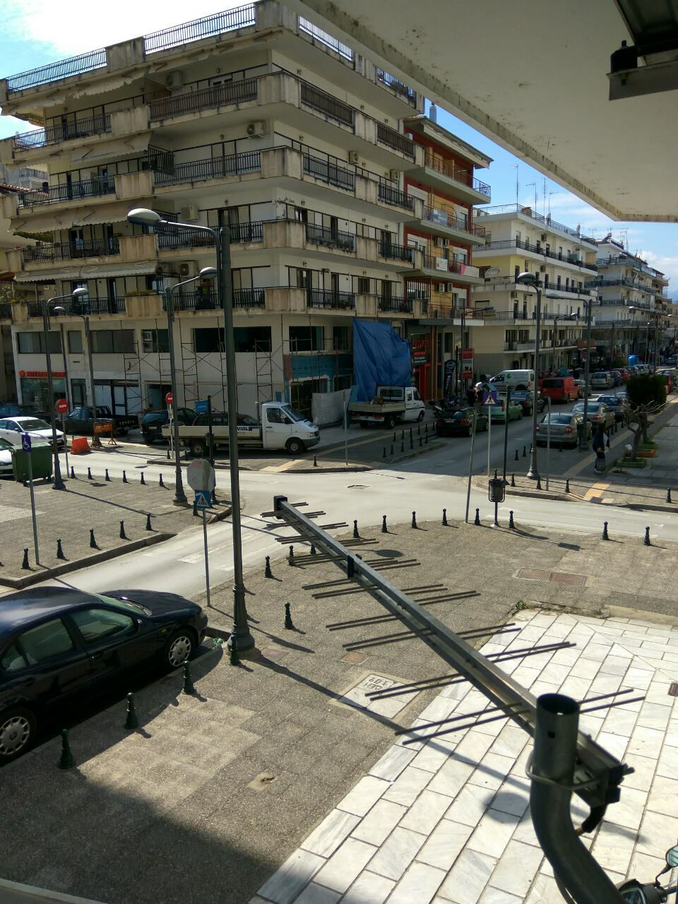 A view of a bustling urban street features a range of buildings, vehicles, and people. Sidewalks are lined with decorative streetlamps and low fences. Construction activity can be seen with a blue tarp covering equipment. The area captures the vibrancy of city life.