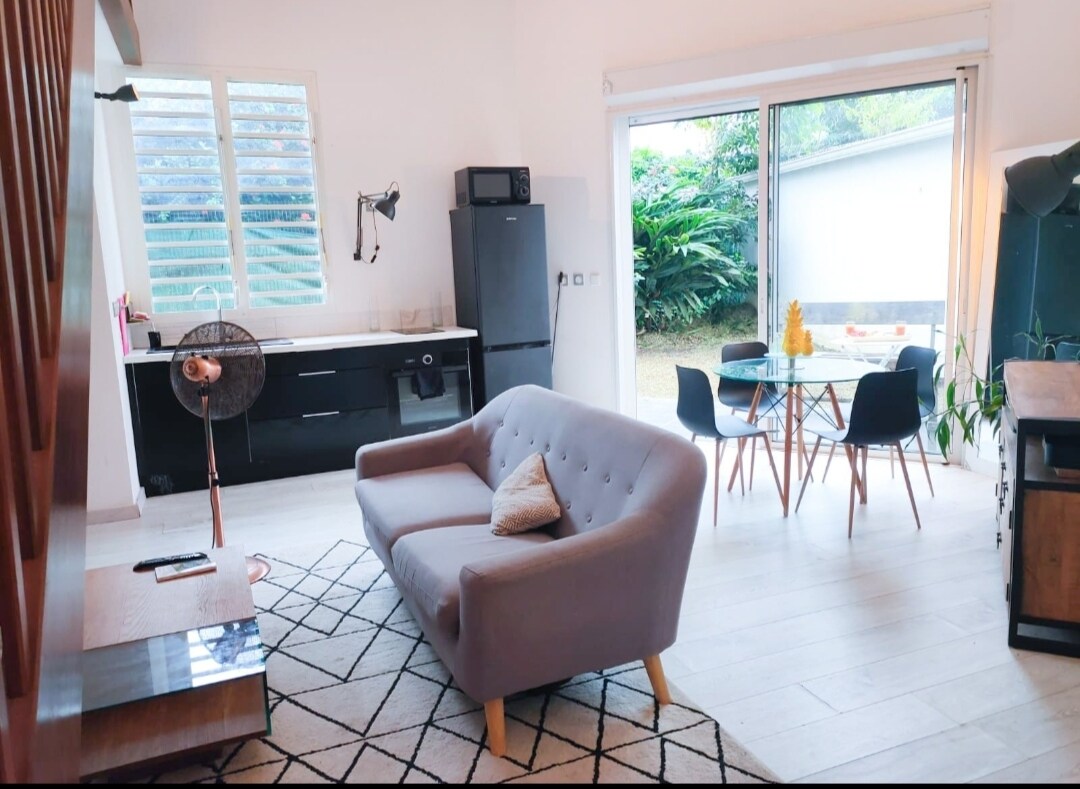 A modern living area showcases a comfortable, light-colored sofa facing a flat-screen TV. A dining table with four chairs is positioned by a large sliding door, allowing views of the garden. The kitchen features sleek black cabinetry and a refrigerator, enhancing the functional space.